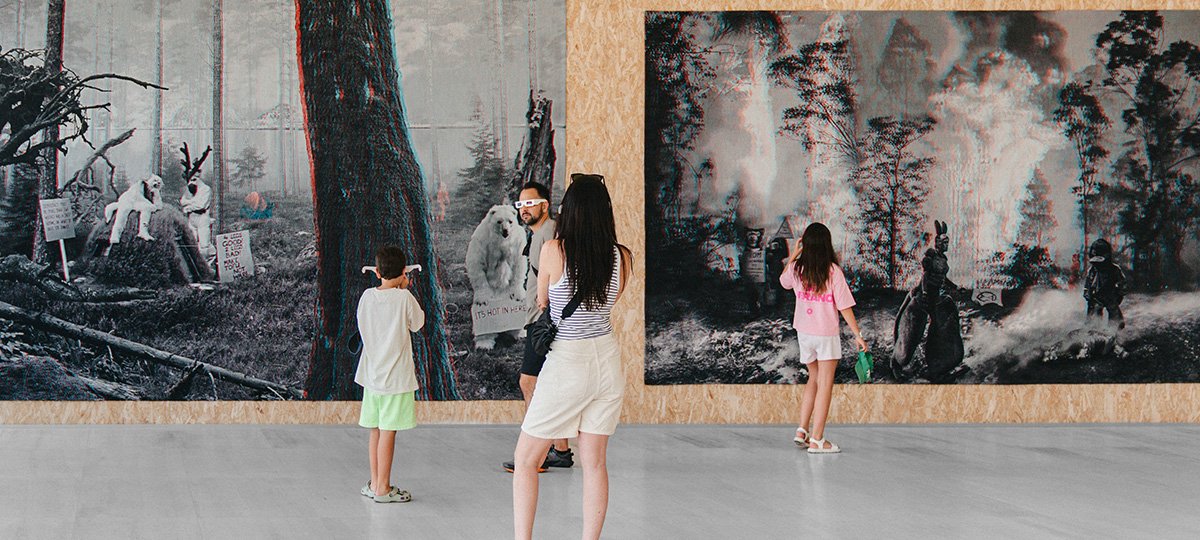 Bambini, bambine e adulti visitano una mostra d’arte contemporanea con grandi installazioni fotografiche presso uno spazio espositivo.